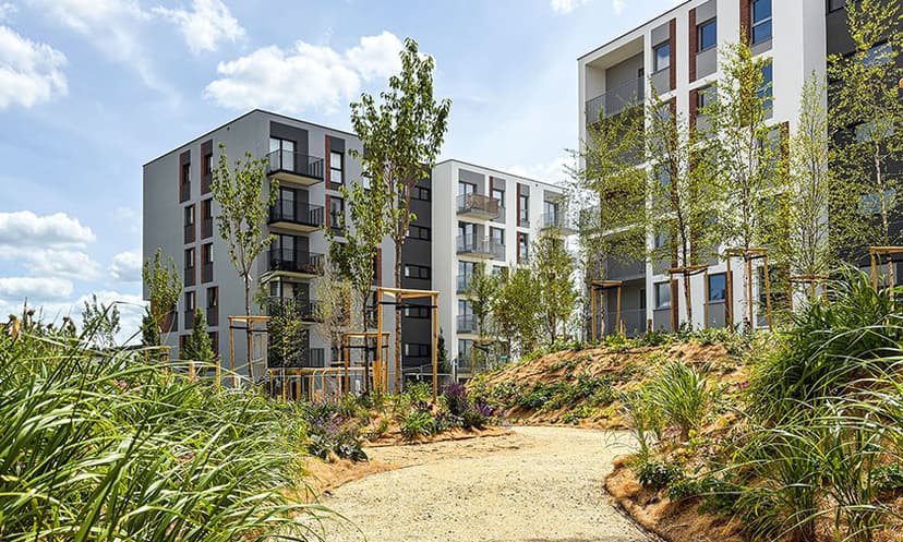 Nowoczesne apartamentowce z balkonami stoją za ogrodami krajobrazowymi i krętą żwirową ścieżką, otoczone zielonymi drzewami i krzewami pod błękitnym niebem - odkryj to spokojne otoczenie z naszą limitowaną ofertą.