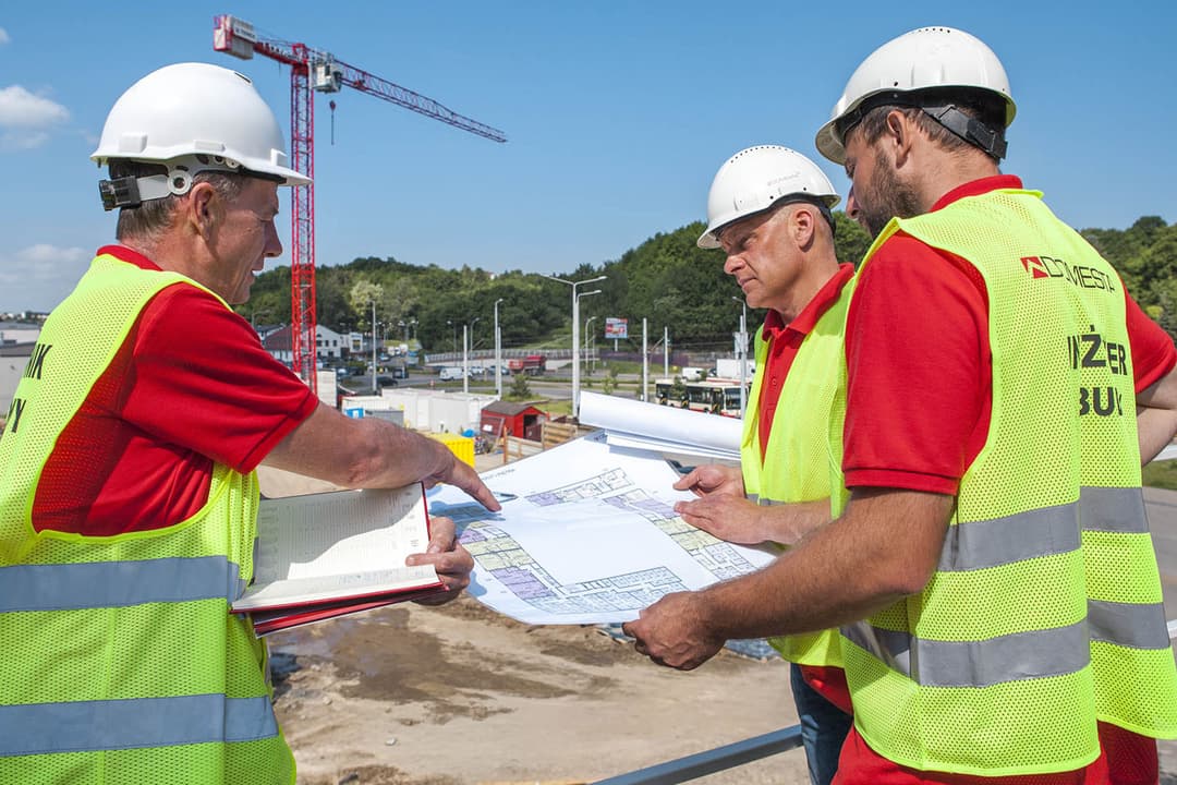 Trzech robotników budowlanych w kaskach i żółtych kamizelkach przegląda plany i notatnik na placu budowy Kariera Domesta, w tle widać dźwig i częściowo zbudowane konstrukcje.
